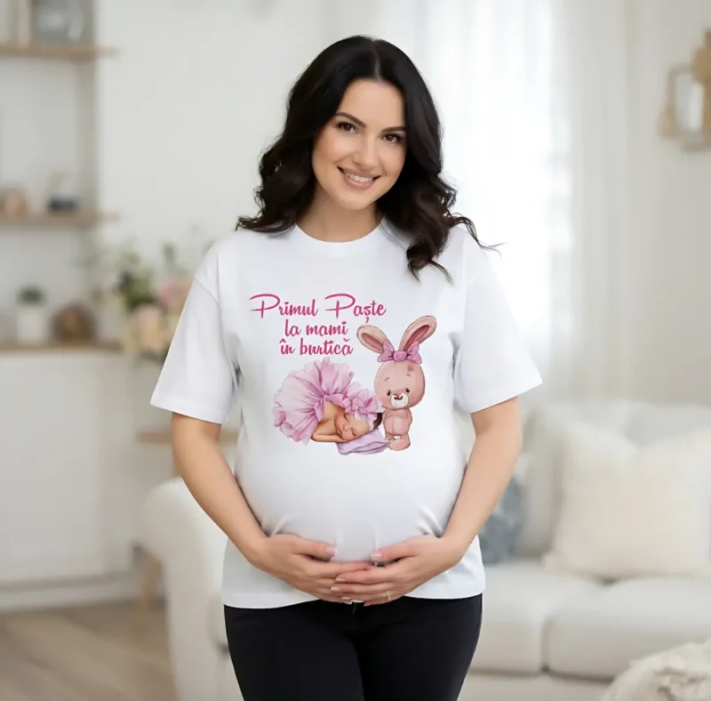 Femeie însărcinată purtând un tricou alb personalizat cu mesajul „Primul Paște la mami în burtică”, ilustrație cu un bebeluș dormind și un iepuraș roz.